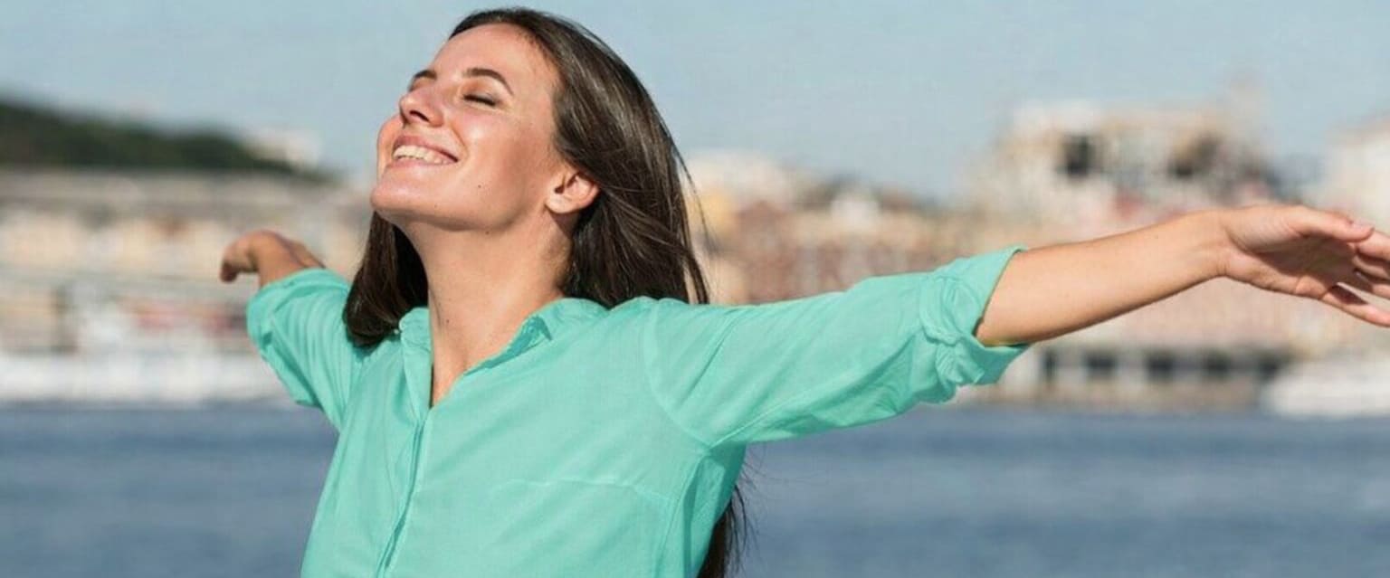 Pessoa ao ar livre, próxima a um corpo de água, com os braços abertos e cabeça inclinada para trás, em postura de relaxamento e bem-estar.