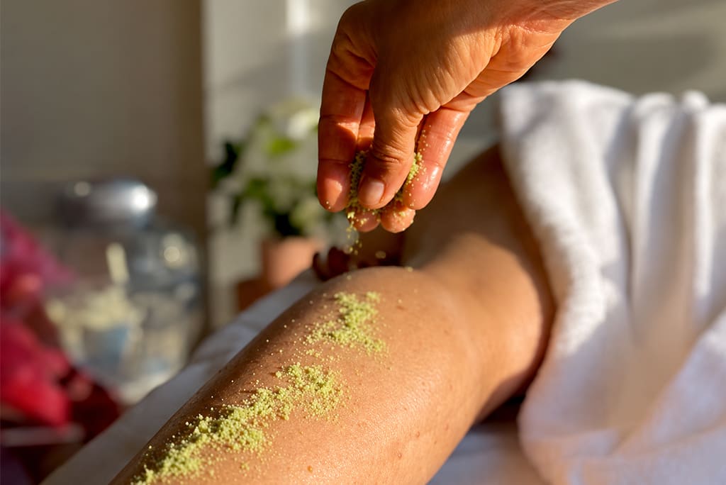 Mão aplicando ervas secas sobre o braço de uma pessoa durante sessão de terapia corporal, em ambiente acolhedor com iluminação suave.